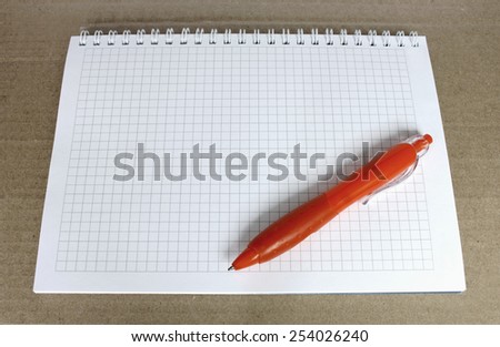 Orange ballpoint pen on the Notepad with a blank page in the box. Notepad on rough brown paper.