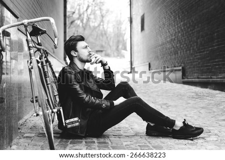 Close up fashion portrait of stylish handsome hipster guy sitting at city park near his fixed bike
