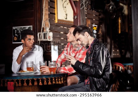 Important call during meeting with friends. Three young men sitting at the bar counter and drinking beer while one of them talks over the phone