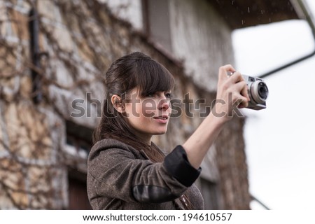 Young attractive woman taking photograph with retro camera in front of cottage in rain