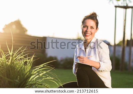 Young happy businesswoman or landscape architect sitting in the garden with her notebook. Young professional smiling at camera and dressed in casual clothes. Strong sunset backlit. Copy space.