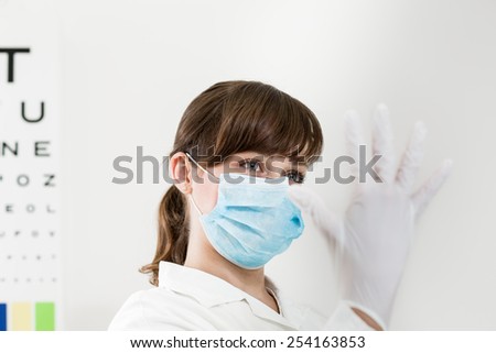 Young female doctor wearing a mask putting on surgical gloves  in the office in front of the eye test chart. Color toned image.