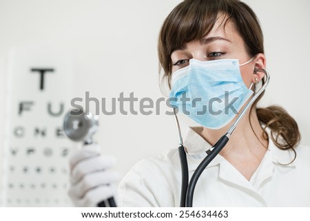 Medical doctor woman carrying stethoscope  in front of the eye test chart. Young female doctor wearing a mask and surgical gloves in the office. Color toned image. Low depth of field. Copy space.