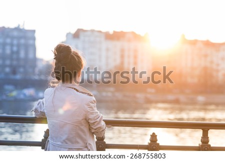 A young woman is looking at the sunset over a river in the city Prague with the old buildings in the background. Soft spring backlit. Color toned image.