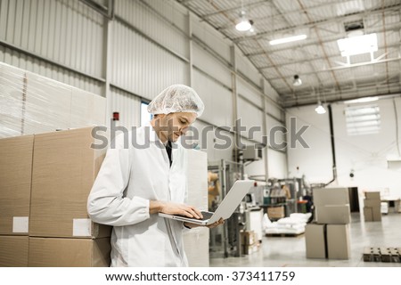Young professional is leaning on the boxes in warehouse for food packaging. Manager is working on the computer in automated production line at modern factory. Color toned image.