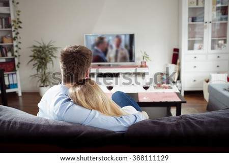 Back view of young couple enjoying themselves and watching tv on the sofa in the living room. Young man and woman relaxing and drinking red wine in the new flat. Man is hugging his girlfriend.