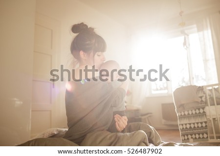 Beautiful young woman with her adorable little son on the bed at home. Young mother kissing her newborn baby. Mother and little boy having good time. Strong sun light from window. Color toned image.