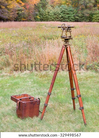 Vintage Brass Surveying Level (Transit, Theodolite) with Compass and aged Brass Patina, on a  wooden Tripod with Storage Case in a field.