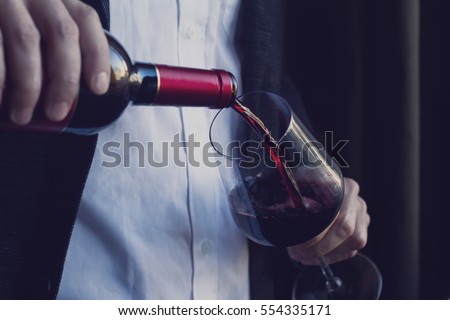 Horizontal close up of Caucasian man in black suit and white shirt pouring red wine into a tall glass in a bar