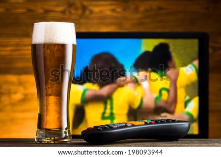 Glass of beer and tv remote, football match in background