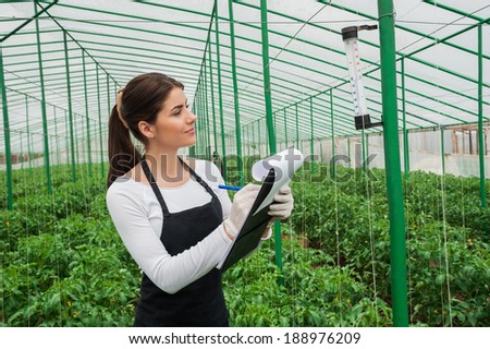 Working in greenhouse. Beautiful woman in uniform writing something in her note pad and looking at the thermometer while standing in greenhouse