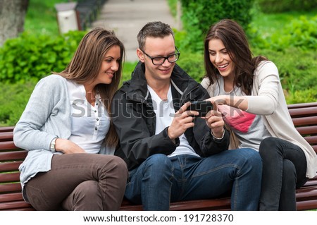 Cheerful group of friends, one man and two women sitting outdoor on a bench in park talking having fun laughing smiling happy watching something on mobile phone .