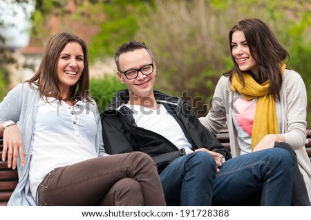 Cheerful group of friends, one man and two women sitting outdoor on a bench in park ,talking having fun, laughing smiling happy.