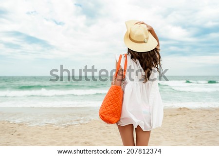 Woman in straw hat and white dress on a tropical beach with orange bag