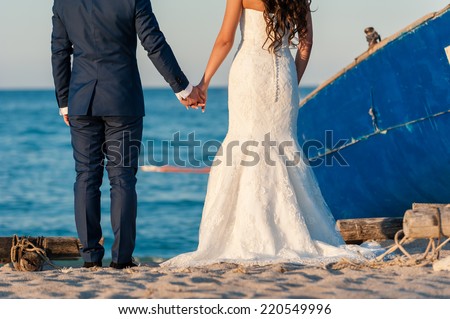 Beautiful stylish newlyweds sitting next to a boat on the beach. Gentle bride and handsome groom by the sea.
