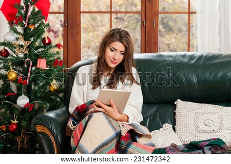 Young woman sitting on couch, alone, in front of christmas tree on living room,using tablet pc.