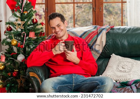 Young man sitting on couch drinking a tea, alone, in front of christmas tree on living room, holding a mug.