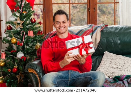 Young man sitting on couch, alone, in front of christmas tree on living room,holding a gift box.