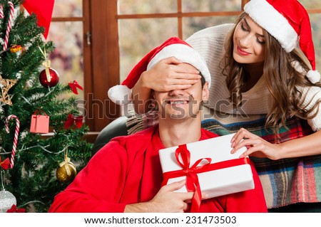 Surprise! Handsome young man sitting on the couch and holding a gift box while her girlfriend standing behind him and covering his eyes with hands and wering santa hat