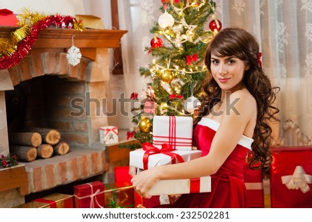 Pretty young woman in red and white christmas dress and long beautiful hair, holding a gift boxes near chimney and Christmas tree over living room.