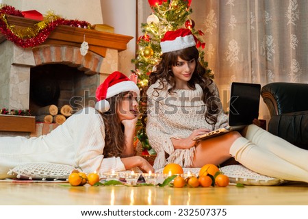 Christmas holiday happy girl friends looking at something on Personal Computer, wearing red new year santa hat, over chimney and christmas tree colorful lights background.