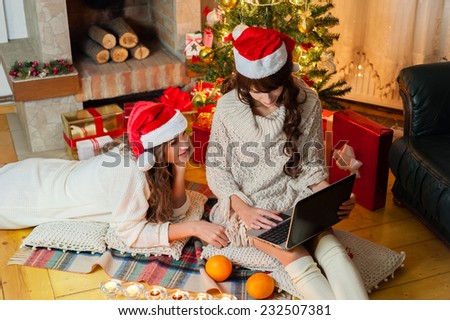 Christmas holiday happy girl friends looking at something on Personal Computer, wearing red new year santa hat, over chimney and christmas tree colorful lights background.