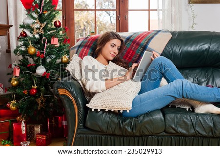 Young woman sitting on couch, alone, in front of christmas tree on living room,using tablet pc.