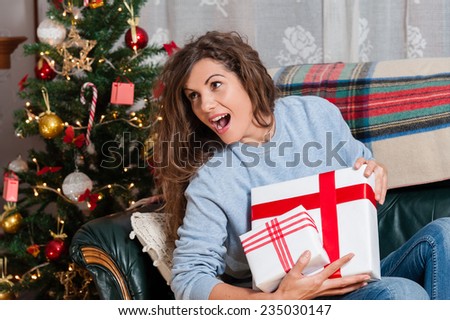 Happy young woman sitting on couch holding christmas present boxes next to christmas tree and fire place
