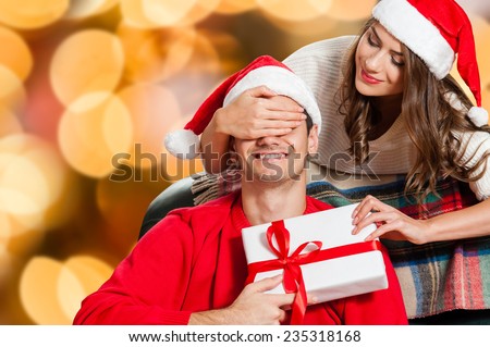 Handsome young man sitting on the couch and holding a gift box while her girlfriend standing behind him and covering his eyes with hands and wearing santa hat christmas colorful lights background