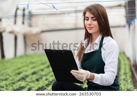 Beautiful young biotechnology woman engineer with clipboard and pen, examining plants for disease in greenhouse with apron and gloves. Greenhouse produce. Food production.