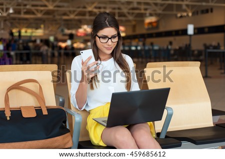 Airport Young female passenger on smart phone and laptop sitting in terminal hall while waiting for her flight. Air travel concept with young casual woman sitting with hand luggage suitcase.