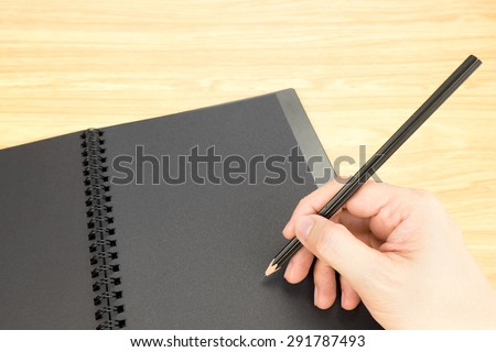 hand holding pencil writing on blank black book with table and business card on wooden table, Mock up for adding your content.