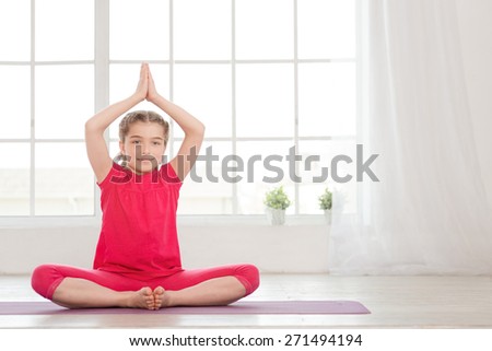 Smiling little girl in lotus position in fitness studio with big windows on background