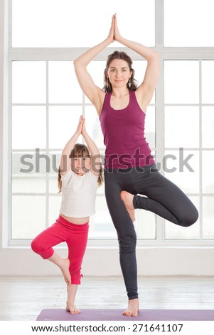 Young mother and daughter doing yoga exercise in fitness studio with big windows on background