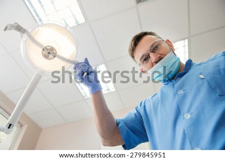 View from the patient side at the dentist. Young dentist approaching a patient and adjusting lamp in dental cabinet