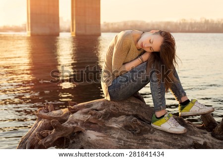 Portrait of beautiful girl with closed eyes sitting on the beam near water on sunset