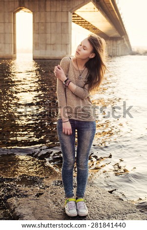 Young sensual woman with closed eyes standing on the stone near water on sunset. Urban landscape