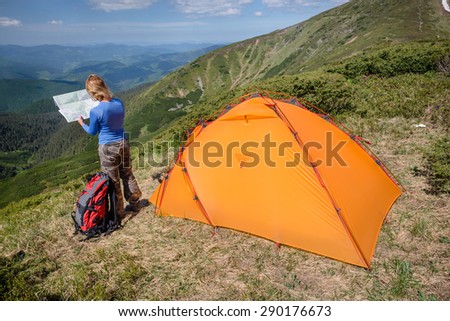 Woman standing near tent and looking at the hike map in the beautiful mountain landscape