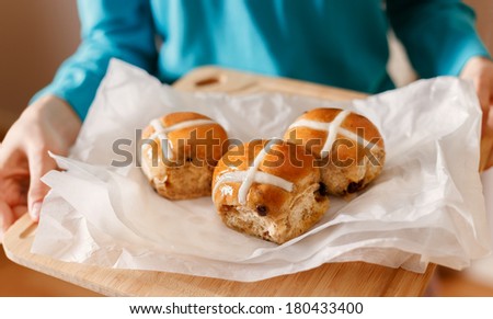 Woman in turquoise clothing holding wooden chopping board with three glazed backed Easter cross buns placed on white baking paper