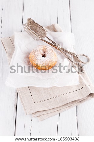 Freshly baked cinnamon sugared doughnuts on baking paper and linen napkin with silver vintage cake tongs on white wooden background. Elegant rustic still life