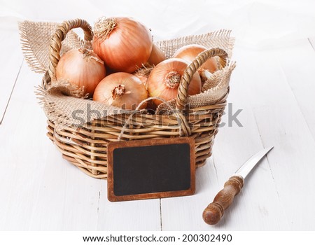 Group of brown onions in a woven basket with tag over white wooden background