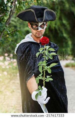 Mystery man in black mask and velvet cape holding a red rose. Selective focus
