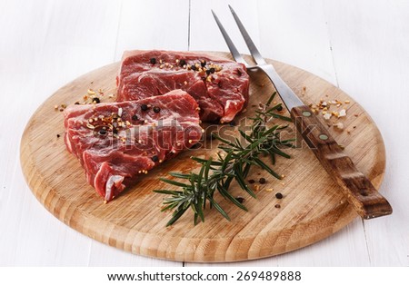 Two pieces of red meat, rosemary, spices  and fork on a round cutting board over white wooden background. Copy space