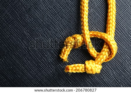 Gold color rope cable with simple knot close up photo hanging down in front of the black color artificial leather background.