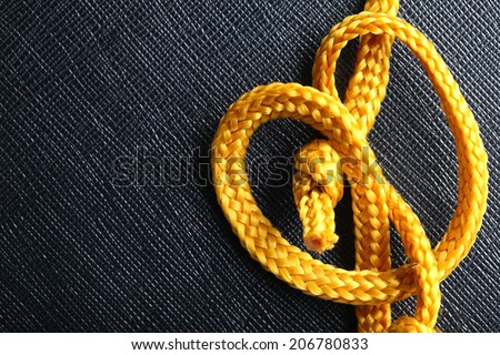 Gold color rope cable with simple knot close up photo hanging down in front of the black color artificial leather background.