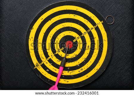 Used and vintage dartboard with arrow in the scene appear a lot of damaged hole from arrow marked put on the black color leather surface as a background represent the entertainment game.