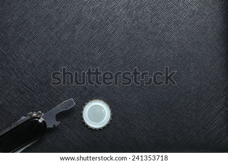 Metallic crown cap beside bottle opener in utility knife set put on black color leather surface background represent the beverage containing equipment