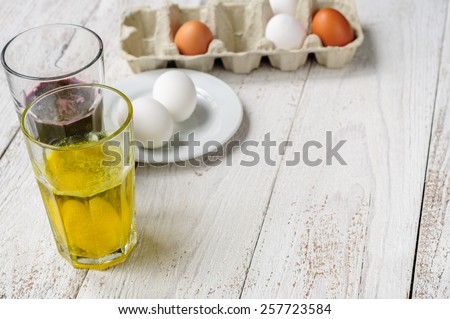 On wooden white shabby background are eggs in carton, eggs on plate and two glasses of food dye with eggs