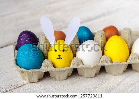 Colored easter eggs in carton with egg-shaped bunny on wooden white shabby background