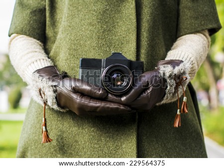 Woman in green pastel coat in autumn background holding a retro camera in hands with leather gloves